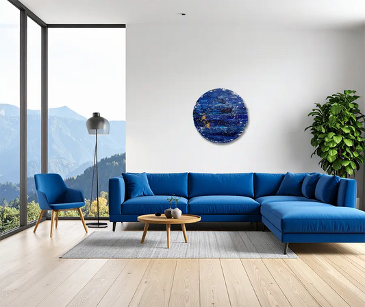 Das runde abstrakte Kunstwerk Lena in blau mit weißen Farbfeldern und goldenen Sprenkeln mit Farbverwischungen und hellen Streifen. An einer hellen Wand hängend mit blauem Sofa.