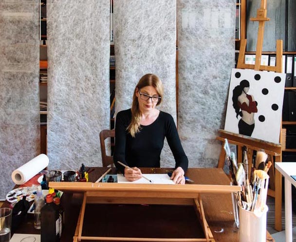 Portrait der Künstlerin im Atelier an Schreibtisch sitzend.
