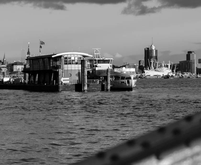 Ein schwarz Weiß Foto der Elbe mit Elbphilharmonie, Hanseatic Trade Center, Fähren und einem Brückenbogen, Detaiansicht der Gebäude auf der anderen Elbseite