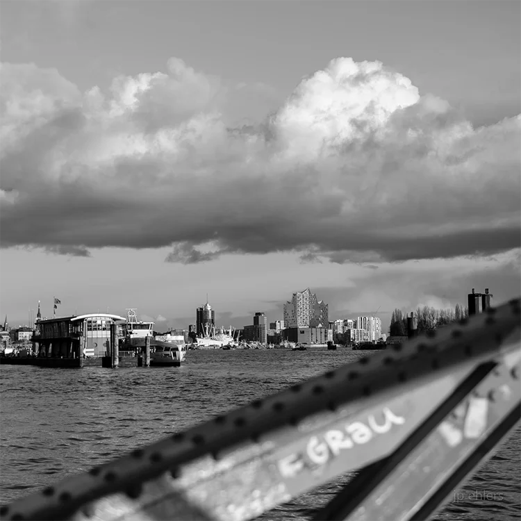 Ein schwarz Weiß Foto der Elbe mit Elbphilharmonie, Hanseatic Trade Center, Fähren und einem Brückenbogen