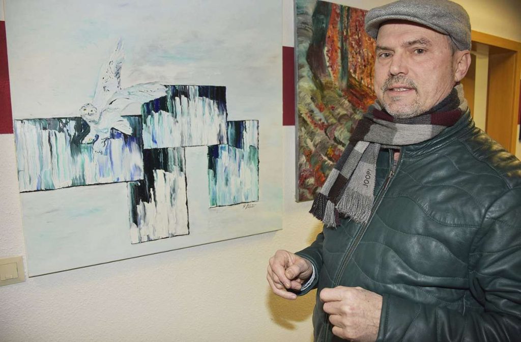 Roger Ziereisen steht vor einem seiner abstrakten Kunstwerke, er trägt eine Mütze und schaut in die Kamera